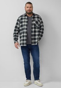 Koszula flanelowa w czarno-białą kratę na szarej koszulce, zestawiona z niebieskimi dżinsami i beżowymi trampkami; swobodny, warstwowy strój.