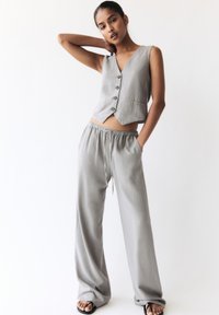 Grauer Leinen-Crop-Top mit Knopfverschluss, kombiniert mit locker sitzenden grauen Hosen mit elastischem Bund und Kordelzug. Sandalen.