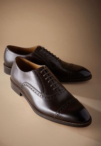 Paire de chaussures Oxford habillées en cuir marron foncé poli avec des détails brogue et des lacets fermés, présentées sur un fond beige.