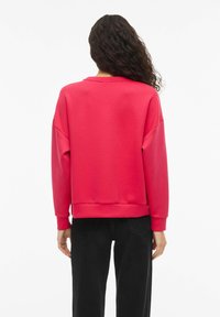 Červená mikina s kulatým výstřihem a spadenými rameny. Vyrobena z měkkého materiálu, má žebrované manžety a lem. Nositelná s černými kalhotami.