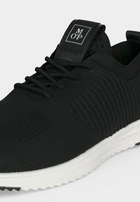 Baskets en maille noire avec lacets noirs, semelle blanche et une étiquette noire sur la languette avec les lettres « M O P » en blanc.