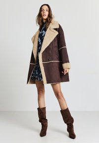 Manteau en shearling marron avec des accents beige, col oversize, poche zippée et coutures contrastantes. Le mannequin porte une robe à motifs et des bottes marron montantes jusqu'aux genoux.