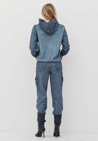 Asmuo su ilgais plaukais, vilkintis mėlyną gobtuvą turinčią striukę, suderintas cargo kelnes ir juodus aukštakulnius batus, stovi susidūręs su paprasta balta siena.