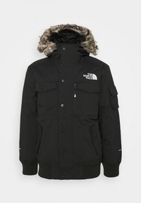 Veste d'hiver noire isolante avec capuche bordée de fourrure, plusieurs poches avant, poignets côtelés et logo The North Face sur la poitrine.