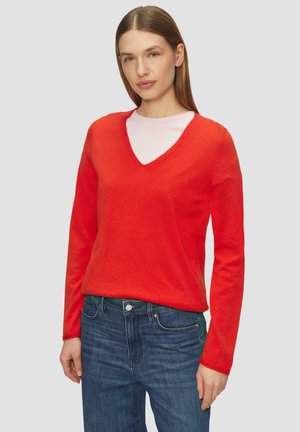Pull en v rouge avec des manches longues, des poignets côtelés et une texture douce, associé à un jeans en denim bleu.