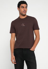Καφέ βαμβακερό t-shirt με μικρό κόκκινο και άσπρο λογότυπο "CK" στο στήθος, με στρογγυλή λαιμόκοψη και κοντά μανίκια.