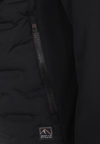 Giacca a vento nera con una texture di tessuto liscia, dotata di una zip impermeabile con un accento rosso e una patch del logo sul rivestimento interno.