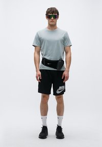 Grå sportt-shirt, svarta shorts med vit logotyp, svart midjeväska, vita strumpor och svarta sneakers, kompletterade med färgglada solglasögon.