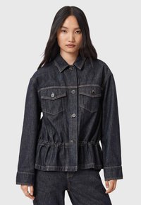 Giovane donna che indossa una giacca di jeans blu scuro con chiusura a bottoni e vita arricciata, abbinata a pantaloni di jeans coordinati, in piedi contro uno sfondo semplice.