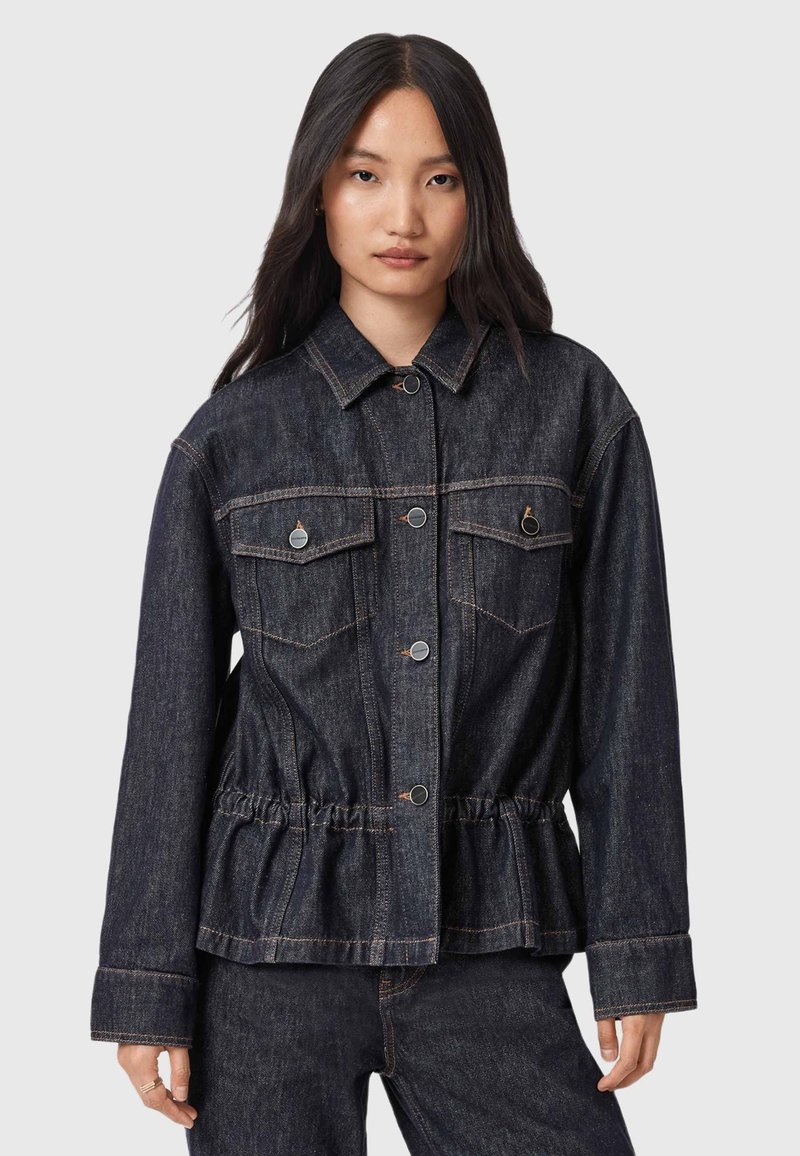 Giovane donna che indossa una giacca di jeans blu scuro con chiusura a bottoni e vita arricciata, abbinata a pantaloni di jeans coordinati, in piedi contro uno sfondo semplice.