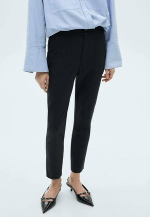 Černé kalhoty na míru se slim fit, hladkou texturou a středním pasem, doplněné světlou modrou košilí a špičatými černými botami.