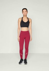 Sujetador deportivo negro, leggings rojos con paneles de malla, ajuste entallado, combinados con zapatillas negras, de pie contra un fondo neutro.