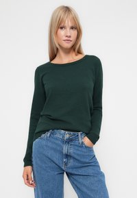 Pull vert foncé avec un motif texturé, col bateau et manches longues, associé à un jean en denim bleu clair. Design décontracté et ajusté.