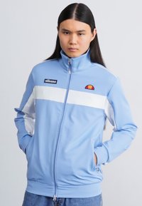 Light blue zip-up jacket made of smooth fabric, featuring white stripes and a small logo. The design includes side pockets and a stand-up collar.