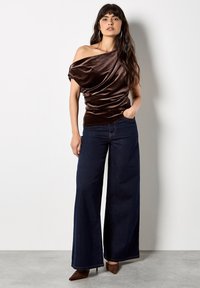 Top de terciopelo marrón sin hombros con diseño drapeado, combinado con pantalones anchos de mezclilla oscura y tacones marrones de punta.