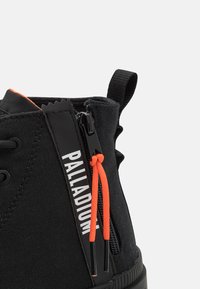 Melnas augstas kedas ar kanvas virsu, kurām ir oranžas akcenta auklas, sānu rāvējslēdzis un kontrastējošs balts logo. Strukturēta gumijas zole.