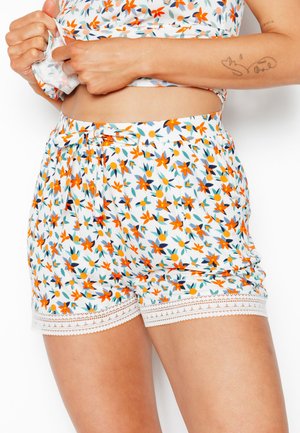 Mains soulevant un haut court à fleurs, portant un short assorti à fleurs avec bordure en dentelle, petit nœud, et un tatouage en dessin linéaire visible sur l'avant-bras.