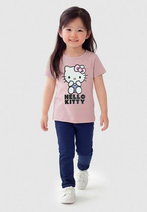 Niña joven caminando, vistiendo una camiseta rosa de Hello Kitty, pantalones azul oscuro y zapatillas blancas, sonriendo sobre un fondo liso.