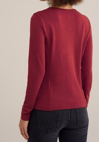 Person with curly hair wearing a fitted long-sleeve maroon top and dark wash jeans, viewed from the back against a beige background.