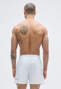 Fehér fürdőnadrág, gumis derékrésszel, melyet hátulról látható férfi visel. Jellegzetes tetoválásminták közé tartozik egy kígyó és egy szív motívum.