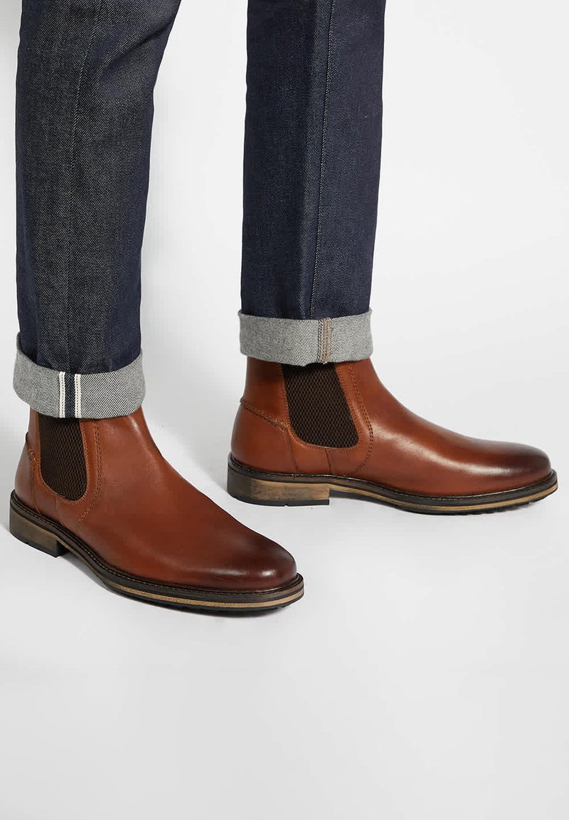 Brown leather Chelsea boots with elastic side panels, a textured surface, and a stacked wooden heel, paired with cuffed dark jeans.