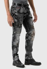 Homme portant un jean à motifs camouflage noir et gris avec un patch "CIPO BAXX" et une ceinture studded noire, assorti de bottes en cuir noires.