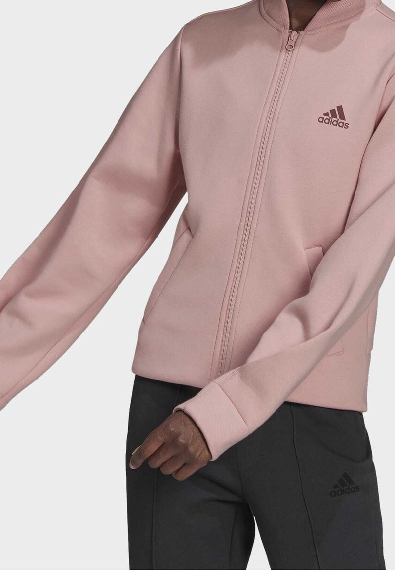 Personne portant une veste zippée rose clair Adidas et un pantalon Adidas noir, le bras droit légèrement tendu vers l'avant.