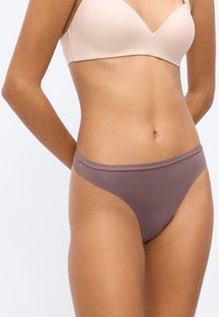 Γυναίκα φορώντας ανοιχτό μπεζ σουτιέν και μοβ αόρατα εσώρουχα, στέκεται μπροστά σε απλό λευκό φόντο.