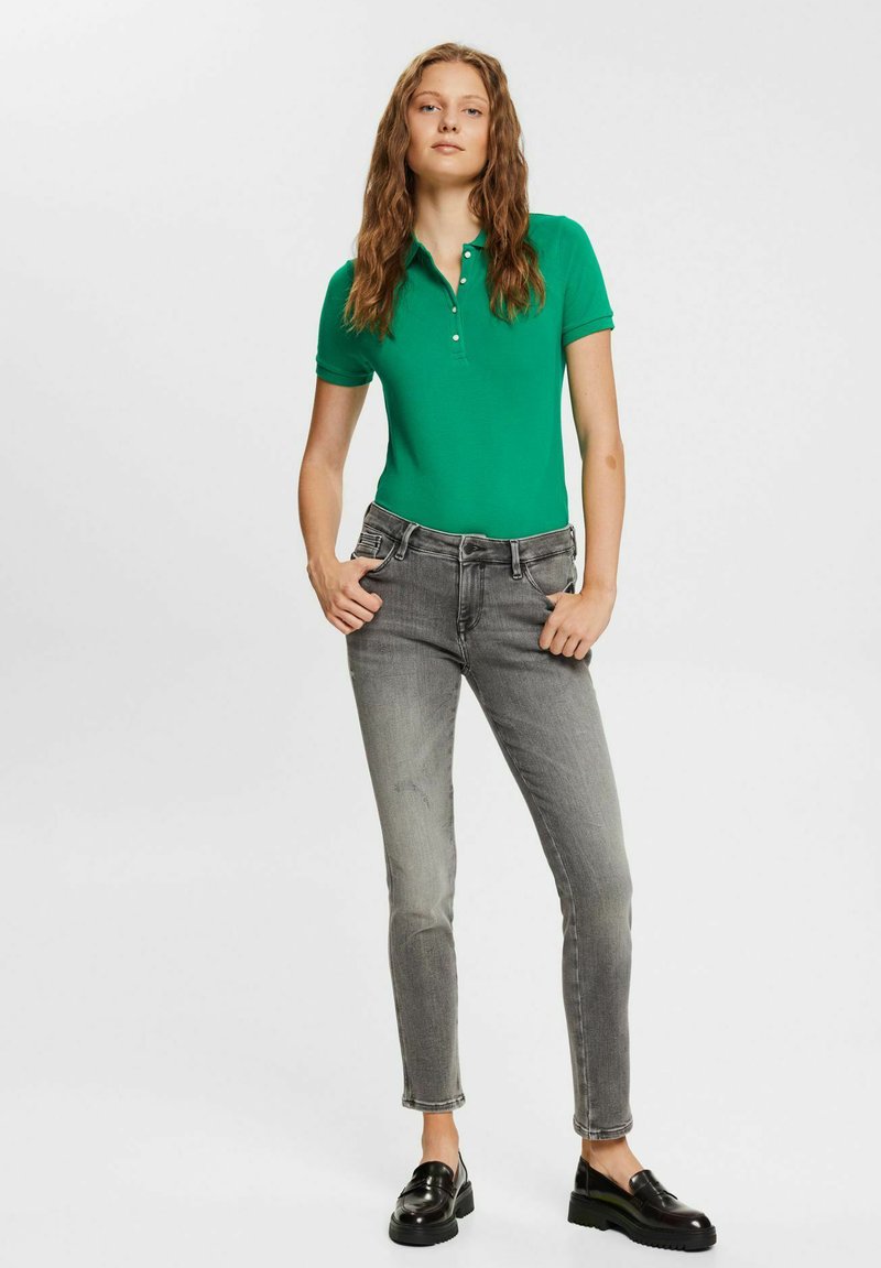 Grünes Baumwoll-Polohemd mit kurzen Ärmeln und einem Drei-Knopf-Verschluss, kombiniert mit grauen Slim-Fit-Jeans und schwarzen Slippern.