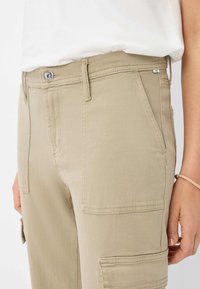 Beige cargobroek met opgestikte zakken, knoop- en ritssluiting, en gestikte details. De stof heeft een gladde textuur.