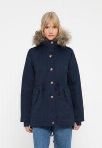 Una donna che indossa un parka invernale blu navy con cappuccio foderato in pelliccia e bottoni frontali, abbinato a jeans di denim blu, frontale.