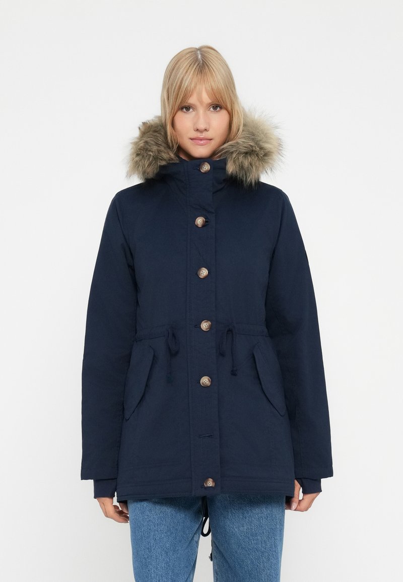 Una donna che indossa un parka invernale blu navy con cappuccio foderato in pelliccia e bottoni frontali, abbinato a jeans di denim blu, frontale.