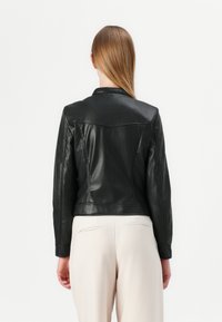 Giacca di pelle nera con un design aderente, caratterizzata da un colletto a punta e cuciture decorative, abbinata a pantaloni chiari.
