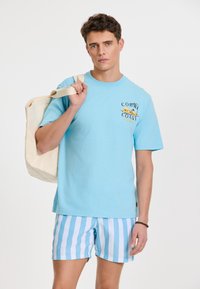 Hellblaues Baumwoll-T-Shirt mit einem grafischen Design, das "Coral Coast" zeigt. Kombiniert mit blauen und weißen gestreiften Shorts und hält eine cremefarbene Tragetasche.