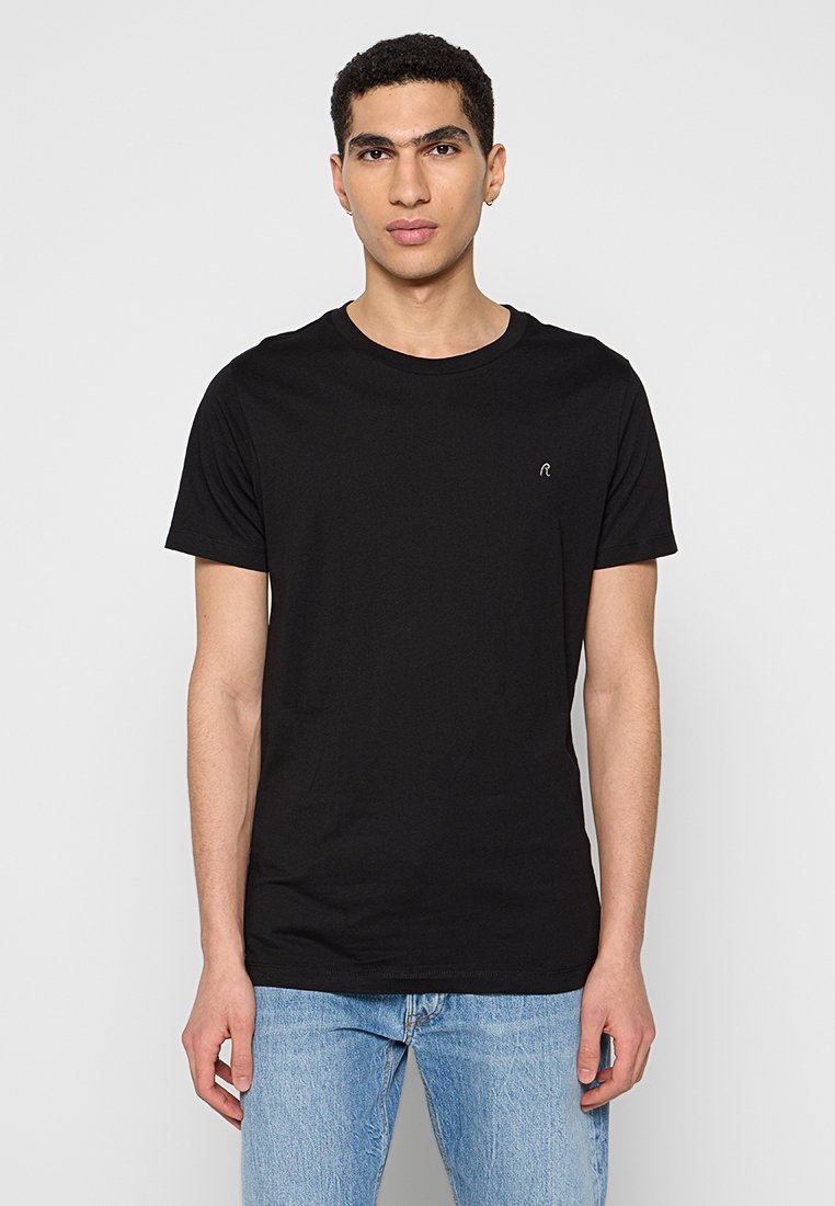 Schwarzes Baumwoll-T-Shirt mit kurzen Ärmeln, Rundhalsausschnitt, das ein kleines gesticktes Logo auf der Brust trägt. Kombiniert mit blauen Jeans.