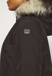 Chaqueta parka negra con un ribete de capucha de piel sintética, bolsillo lateral y un parche de marca en el hombro. Textura suave y material duradero.