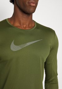 Mann trägt ein olivgrünes Langarmhemd mit einem großen, strukturierten weißen Nike-Swoosh-Logo auf der Brust.