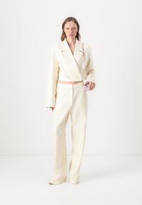Cropped double-breasted blazer and wide-leg trousers in off-white textured fabric. Model holds a small pink bag.