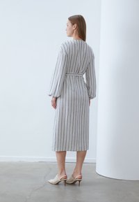 Gestreiftes Midikleid in Hellgrau mit dunkleren Streifen, mit langen Ärmeln, einem Taillengürtel und lockerem Schnitt. Kombiniert mit beigen offenen Schuhen.