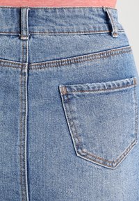 Jeans en denim bleu taille haute avec un délavage, dotés de surpiqûres orange et d'une seule poche arrière de forme rectangulaire.