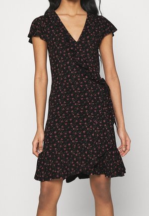 Czarna sukienka kopertowa z małym różowym kwiatowym wzorem, krótkie falbankowe rękawy, dekolt w serek i marszczony dół, noszona przez stojącą osobę.