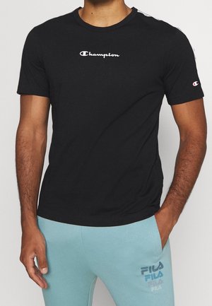 Man draagt een zwart Champion-T-shirt en lichtblauwe FILA joggingbroek, staat met één hand in de zak tegen een effen achtergrond.