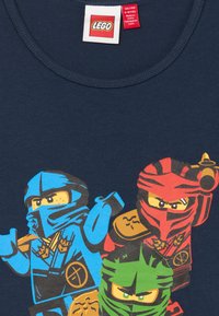 Triá LEGO ninja postavičiek v modrých, červených a zelených maskách na tmavomodrom tričku veľkosti 9-10 rokov od značky LEGO.