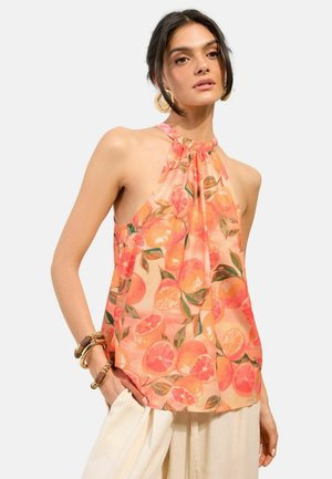 Vrouw met een mouwloze haltertop met sinaasappelprint, crèmekleurige broek met wijde pijpen, gouden oorringen en meerdere armbanden, handen in de zakken.