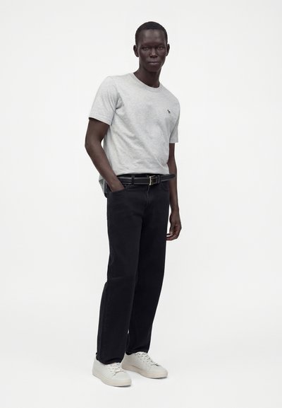 Homme portant un t-shirt gris clair, pantalon noir avec ceinture noire et baskets blanches, debout avec une main dans la poche sur un fond blanc uni.