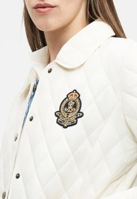 Κοντινό πλάνο ατόμου που φοράει λευκό καπιτονέ μπουφάν με γιακά και κεντημένη κονκάρδα με το έμβλημα Ralph Lauren στο στήθος.
