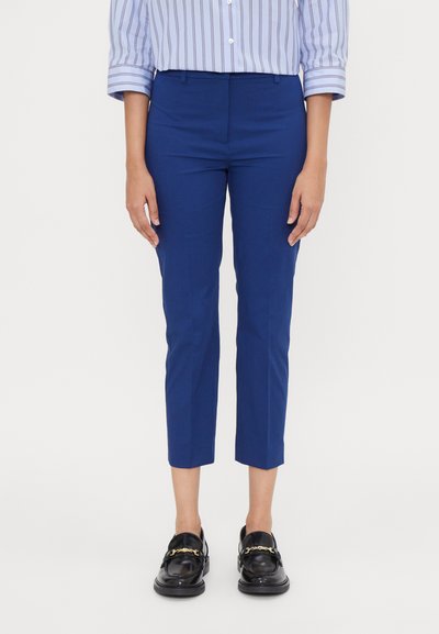 Pantalons de costume bleus coupés dans un tissu lisse, présentant une coupe ajustée et des poches avant discrètes. Portés avec des mocassins noirs.