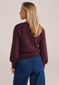 Pull en tricot bordeaux avec texture côtelée, manches ballon et taille cintrée, associé à un pantalon en denim bleu à jambes larges avec poches arrière.