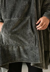 Grauer gestrickter Poncho mit strukturierter Oberfläche, ausgestattet mit einer großen Fronttasche und einem asymmetrischen Saum, der ein darunter liegendes schwarzes Kleidungsstück teilweise verdeckt.