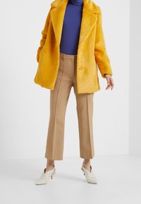 Abrigo de piel sintética amarilla con cuello y cierre de botones; combinado con pantalones beige ajustados y zapatos de tacón bajo blancos sin cordones.